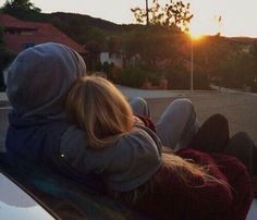 two people sitting on top of a car in front of the sun with their arms around each other