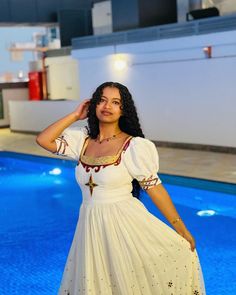 a woman standing in front of a swimming pool wearing a white dress with red trimmings