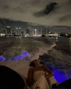 a woman laying on top of a boat in the water near a city at night