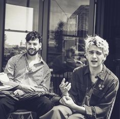 two men sitting next to each other in front of a glass window and one is holding his hand out