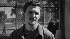 a man with a mustache standing in front of a chain link fence and looking at the camera