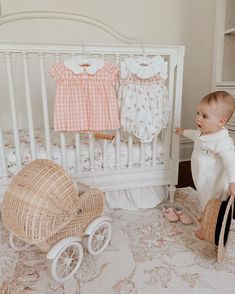 a baby standing in front of a crib with clothes hanging on the wall next to it