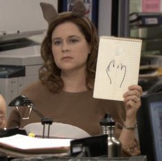 a woman sitting at a desk holding up a piece of paper with a face drawn on it