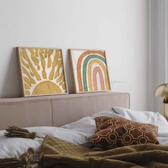 two paintings on the wall above a bed with white linens and brown throw pillows