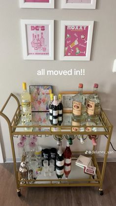 a gold bar cart with liquor bottles and glasses on it in front of two framed pictures