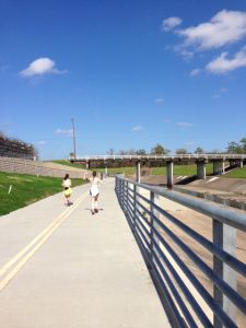 two people walking down a sidewalk next to a bridge and grass covered field with trees