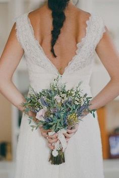 a woman in a wedding dress holding a bouquet of flowers and greenery with her back to the camera