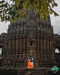 a man and woman standing in front of a large stone structure with an orange skirt