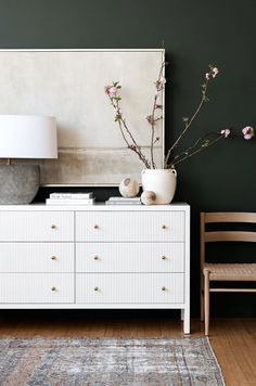 a white dresser sitting next to a lamp and a vase with flowers on top of it