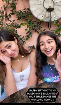 two young women sitting next to each other in front of a wall with plants on it