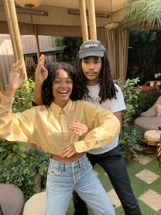 two people standing next to each other in front of some plants and trees with one person holding his arms up