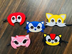 five masks with different colored faces on a wooden surface, one has eyes and the other has ears