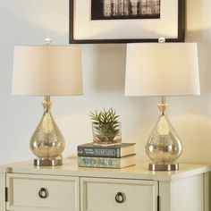 two lamps sitting on top of a white dresser next to a framed picture and books