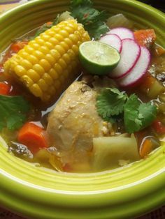 a green bowl filled with chicken, vegetables and corn on the cob next to sliced cucumbers