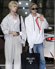 two young men standing next to each other in front of a white car and one holding a black prada bag