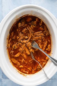 a white bowl filled with meat and sauce on top of a blue table cloth next to a fork