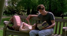 a man laying on top of a wooden bench next to a woman holding a book