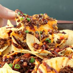 a hand holding a tortilla chip filled with chili cheese and ground beef topped with green onions