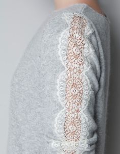 the back of a woman's grey sweater with lace on sleeves and cuffs, standing against a wall