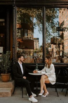 a man and woman sitting at a table in front of a store window talking to each other