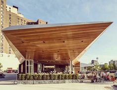 a large wooden structure with people sitting at tables under it in front of tall buildings