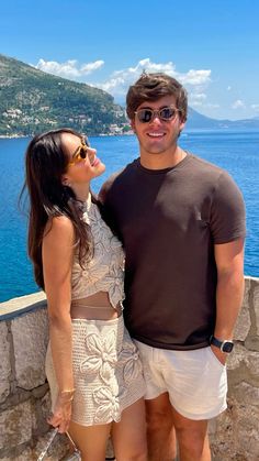 a man and woman standing next to each other on a stone wall near the ocean