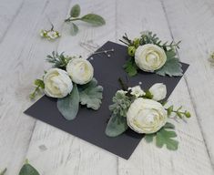 three white flowers are placed on top of a black piece of paper with greenery