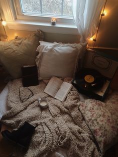 an unmade bed with books, tea cup and other items on it in front of a window