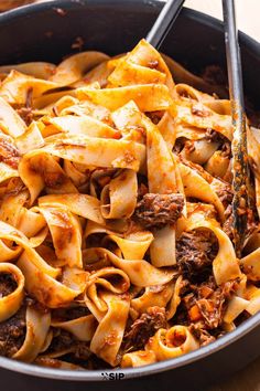 a skillet filled with pasta and meat on top of a wooden table next to a fork