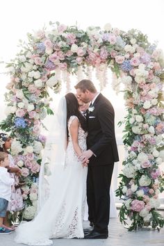 a bride and groom kissing under a floral arch at the end of their wedding ceremony