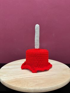 a crocheted red cake with a white top sitting on a wooden table in front of a pink wall