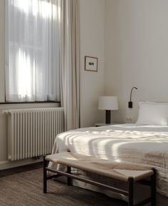 a white bed sitting next to a window with curtains on the windowsill and a bench in front of it