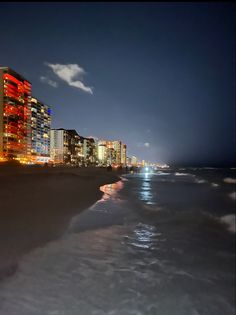 the city is lit up at night on the beach by the water's edge