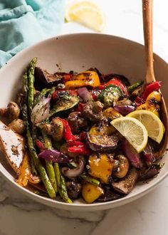 a white bowl filled with mushrooms, asparagus and other vegetables next to a wooden spoon