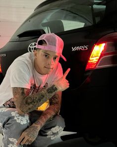 a man with tattoos sitting on the ground next to a black car and giving thumbs up