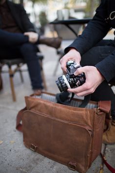 a person holding a camera in their hand while sitting on the ground next to a brown bag