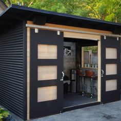 a small black shed with sliding doors on the inside and outside, in front of some trees