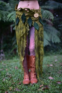 a woman wearing a green skirt and boots with leaves on her legs, standing in the grass