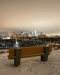 a wooden bench sitting on top of a snow covered slope next to a city skyline