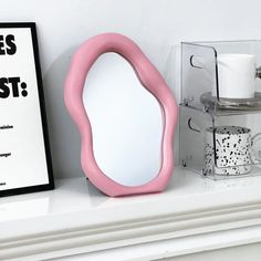 a pink mirror sitting on top of a white mantle next to a cup and mug