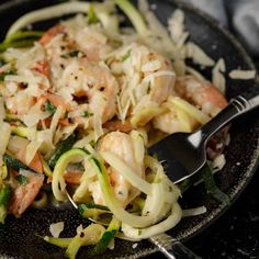 a black plate topped with pasta and shrimp next to a fork on top of a table