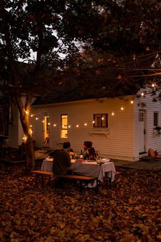 two people sitting at a table in front of a house with lights strung from the roof