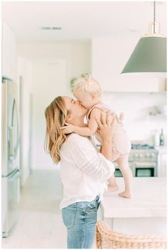 a woman holding a baby up to her face while standing in front of a kitchen counter