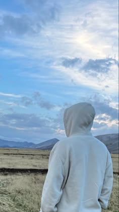 a man in a white hoodie is standing on a grassy field with mountains in the background