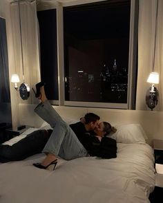 two people laying on a bed in front of a window with the city lights behind them