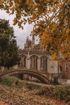 an old building with a bridge in the foreground and autumn leaves on the ground