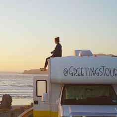 a woman sitting on the roof of a white truck next to the ocean at sunset