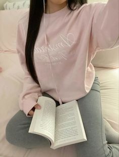 a woman sitting on top of a bed holding an open book and listening to headphones