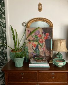 a wooden dresser topped with vases filled with flowers next to a mirror and lamp