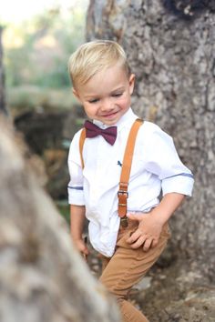 a young boy wearing a bow tie and suspenders standing in front of a tree